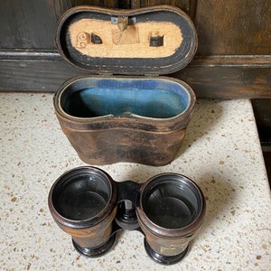 May include: A pair of vintage binoculars with a leather case. The binoculars have black lenses and a brown leather body. The case is brown leather with a blue lining.