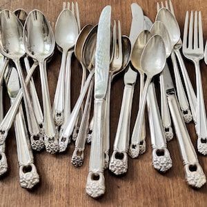 May include: A collection of vintage silverware, including forks, spoons, and knives, arranged on a wooden surface. The silverware has ornate floral detailing on the handles and a shiny, reflective finish. The arrangement suggests a set for dining.