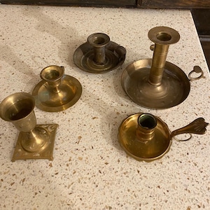 May include: Four antique brass candle holders. Two of the holders have a round base and a single candle holder. One holder has a square base and a single candle holder. The fourth holder has a round base and a single candle holder with a handle.