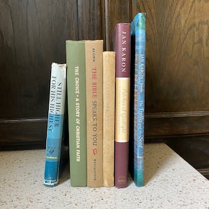 May include: A stack of six paperback books with different titles and authors. The books are arranged vertically with the spines facing out. The books are all different colours and have different designs on the covers. The books are on a light-coloured surface.