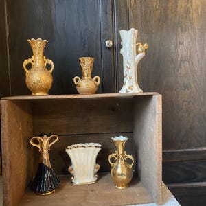 May include: A collection of decorative vases in various styles and colors. The vases are displayed on a wooden shelf. The vases include gold, black, and white colors. Some vases have handles, while others have unique shapes and designs.