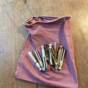 May include: A dusty rose-colored canvas clothespin bag with a wire hanger and several wooden clothespins. The bag is open, and the clothespins are visible inside and outside the bag. The background is a wooden surface.
