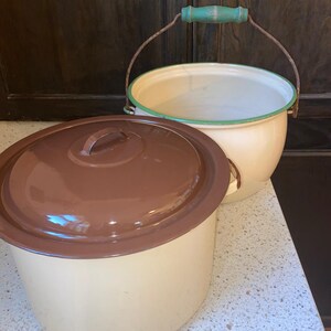 May include: Two vintage enamelware pots with lids. The larger pot has a brown lid and a cream-colored body. The smaller pot has a green rim and a wooden handle.