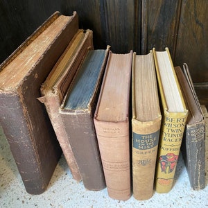 May include: A collection of antique books with worn, textured covers in shades of brown, tan, and blue. Titles include "From Death to Morning" by Thomas Wolfe and "The House in the Mist". The books are arranged on a light-colored surface against a dark wood background.