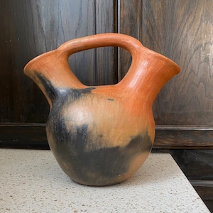 Puede incluir: Una jarra de cerámica artesanal de dos picos con un esmalte negro y naranja. La jarra tiene un cuerpo redondeado y una sola asa.