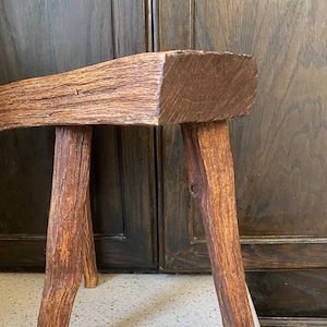 May include: A rustic wooden stool with three legs. The stool is made of rough-hewn wood and has a natural brown finish.