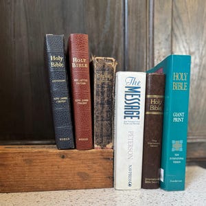 Könnte beinhalten: Eine Sammlung von Hardcover-Büchern, darunter Bibeln und ein Wörterbuch, auf einer kleinen Holzkiste angeordnet. Die Bücher sind in verschiedenen Farben gehalten, mit Titeln wie "Holy Bible" und "The Message".