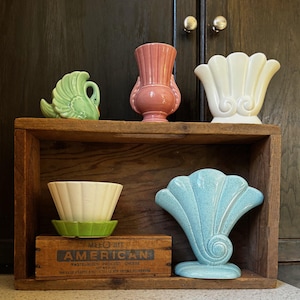 Könnte beinhalten: Ein Holzregal zeigt eine Sammlung von Vintage-Keramikvasen und dekorativen Stücken. Zu den Artikeln gehören ein grüner Schwan, eine rosa Vase, eine weiße muschelförmige Vase, eine hellblaue fächerförmige Vase und eine weiße Schale mit grünem Sockel. Eine Holzkiste mit dem Wort "AMERICAN" ist ebenfalls vorhanden.