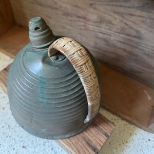 May include: Antique bronze-colored metal kettle with a woven handle. The kettle has a rounded body with horizontal ridges and a small spout. The handle is made of woven light brown material. The kettle is resting on a wooden surface.