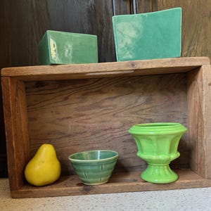 Op de afbeelding: Een houten kist toont een verzameling vintage items. Twee groene rechthoekige keramische stukken liggen bovenop. Binnenin zijn een gele peer, een kleine groene kom en een groene vaas gerangschikt.