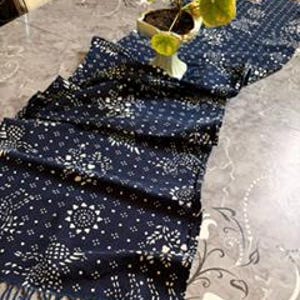 May include: A dark blue table runner featuring a repeating white floral and geometric pattern with a fringed edge. The fabric is draped in folds, and a small white pot containing a green plant with round leaves sits on the runner.