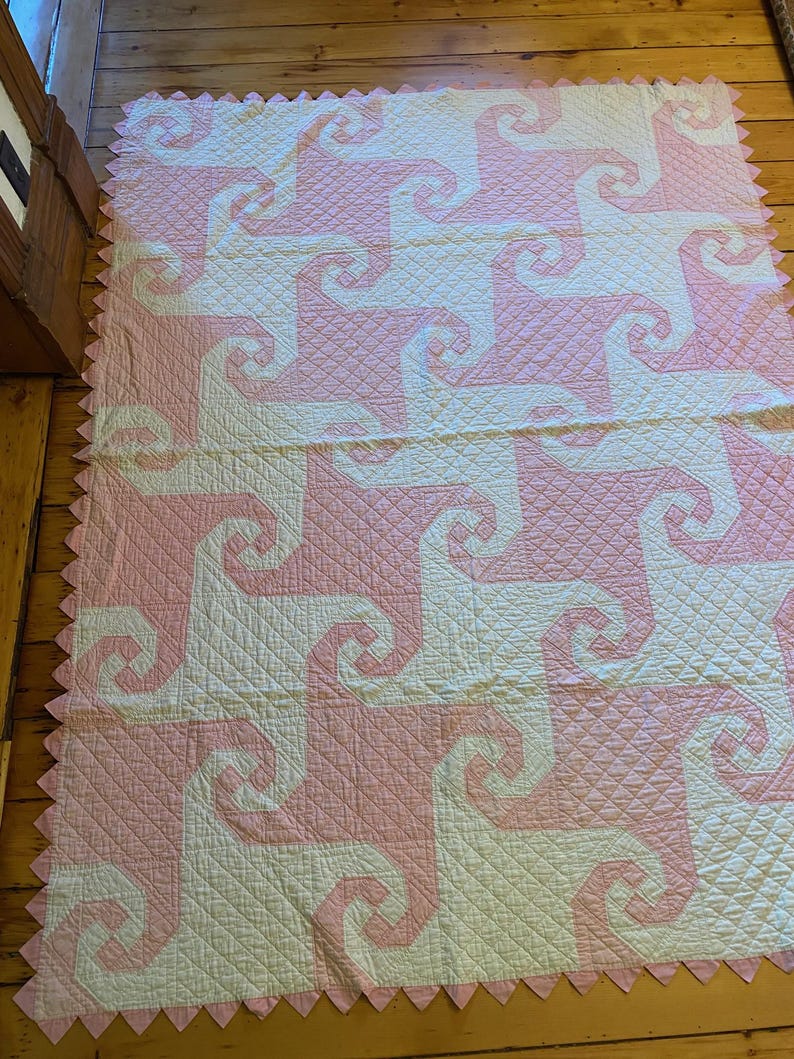 May include: A rectangular quilt featuring a repeating geometric pattern of interlocking pink and white spiral shapes. The fabric surface displays a fine diamond-shaped quilted texture throughout, and the edges are finished with a pink sawtooth border.