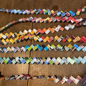 May include: Several strands of handmade paper bead jewelry are displayed on a wooden surface. The beads are made from colorful recycled paper, arranged in a zig-zag pattern. The color palette includes blues, oranges, yellows, and greens.