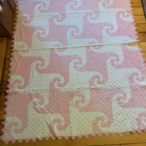May include: A rectangular quilt featuring a repeating spiral pattern in alternating light pink and cream fabric. The surface has a diamond-shaped quilted texture throughout, and the edges are finished with a pink sawtooth border.