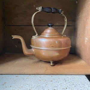 May include: A vintage copper tea kettle with a black wooden handle and brass accents. The kettle has a round body and a spout on the side. It sits on three small feet.