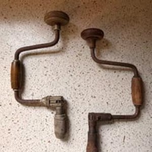 May include: Two antique hand drills with wooden handles and metal frames. The drills have a crank design with a wooden knob at the top and a chuck for holding drill bits. The metal parts show signs of age and wear.