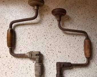 May include: Two antique hand drills with wooden handles and metal frames. The drills have a crank design with a wooden knob at the top and a chuck for holding drill bits. The metal parts show signs of age and wear.