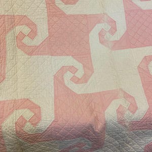 May include: A quilted textile featuring a repeating geometric pattern of interlocking white and light pink spiral shapes. The fabric surface displays a consistent grid of fine, diagonal stitched lines across the entire design.