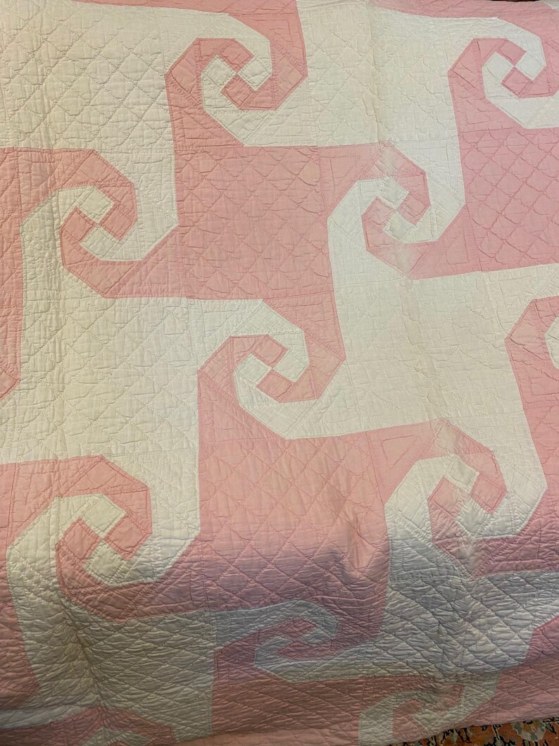 May include: A quilted textile featuring a repeating geometric pattern of interlocking white and light pink spiral shapes. The fabric surface displays a consistent grid of fine, diagonal stitched lines across the entire design.
