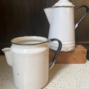 Puede incluir: Dos cafeteras de esmalte blanco con asas y ribetes negros. Una cafetera es más alta con tapa, la otra es más baja. Ambas tienen boquillas para verter. Las cafeteras están sobre una superficie clara con un bloque de madera en el fondo.