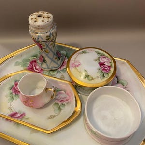 Op de afbeelding: Een set porseleinen toiletartikelen bestaande uit een groot dienblad, een klein kopje met oor, een ronde houder met deksel en een hoge strooier, allemaal voorzien van handgeschilderde roze rozenmotieven, groen blad en goudkleurige randen.