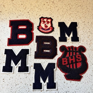 Op de afbeelding: Assortiment van varsity letter patches in marineblauw en rood. Bevat de letters "B" en "M", een schildembleem en een muzikale lier met de tekst "BHS". De patches liggen op een licht gespikkeld oppervlak.