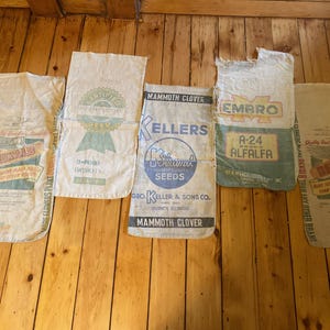 May include: Five rectangular fabric seed sacks laid out in a row. The sacks feature various printed logos and text including DEKALB, CERTIFIED SEED, MAMMOTH CLOVER, KELLERS, EMBRO, and A-24 BRAND ALFALFA. The bags are off-white with green, blue, and yellow ink.