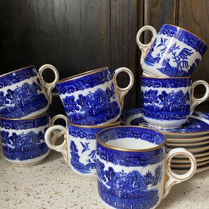 Puede incluir: Un conjunto de tazas de cerámica azules y blancas con ribete dorado. Las tazas presentan un diseño clásico de Willow. Algunas tazas están apiladas y otras sobre platillos. Las tazas tienen asas blancas.