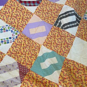 May include: Close-up of a patchwork quilt with a variety of fabric patterns. The quilt features diamond-shaped blocks with floral, plaid, and striped designs in shades of yellow, red, blue, and purple.