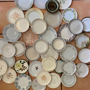 Może przedstawiać: Duża kolekcja różnorodnych talerzy ceramicznych i szklanych ułożonych w grupę. Talerze mają różne wzory, w tym motywy kwiatowe, kłosy pszenicy, złote obwódki, jednolite kolory oraz teksturowane krawędzie w kolorach białym, kremowym, brązowym i zielonym.