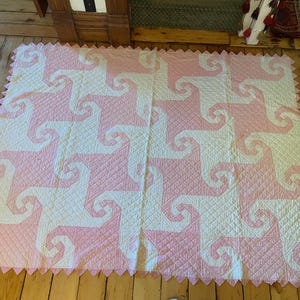 May include: A rectangular quilt featuring a repeating pattern of interlocking white and light pink spiral shapes. The fabric has a quilted diamond texture throughout, and the edges are finished with a decorative pink sawtooth border.