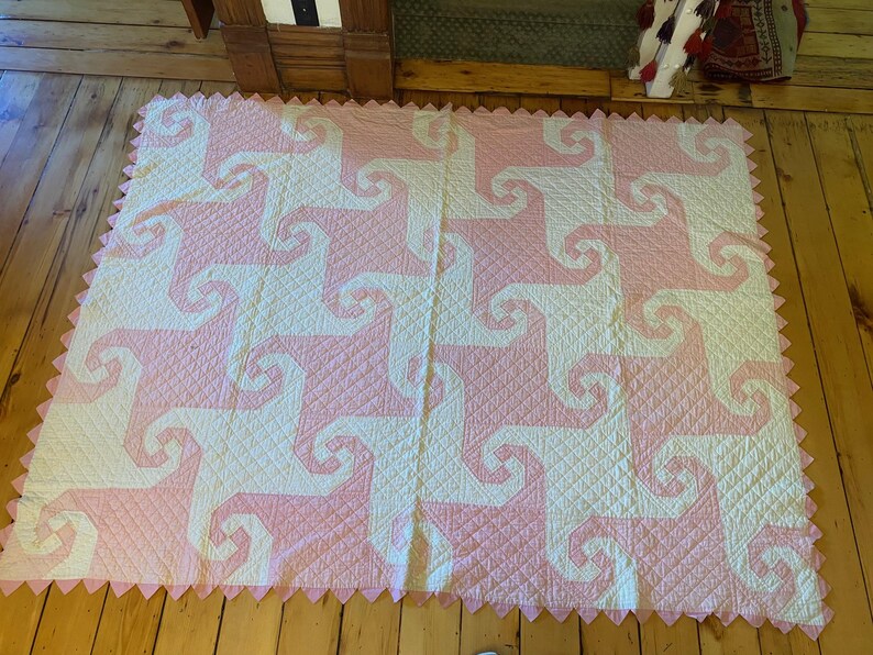 May include: A rectangular quilt featuring a repeating pattern of interlocking white and light pink spiral shapes. The fabric has a quilted diamond texture throughout, and the edges are finished with a decorative pink sawtooth border.