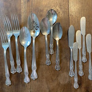May include: A collection of vintage silverware, including forks, spoons, and butter knives, arranged on a wooden surface. The silverware has ornate handle detailing and a polished, reflective finish, suggesting a classic or antique aesthetic.