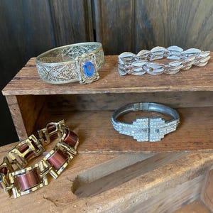 May include: Four vintage bracelets on a wooden surface. One bracelet is silver with a blue stone, one is silver with a geometric design, one is silver with a leaf design, and one is gold with brown rectangular links.