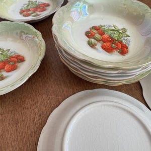 Puede incluir: Colección de cuencos y platos de cerámica vintage. Los cuencos tienen un borde festoneado y un acabado nacarado con un diseño de fresas. Los platos son blancos con un esmalte craquelado.