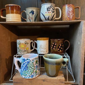 May include: A collection of eight ceramic mugs with various designs and colors. The mugs are displayed on a wooden shelf and a wire rack. The mugs feature floral, abstract, and geometric patterns, with earth-tone and vibrant color schemes.