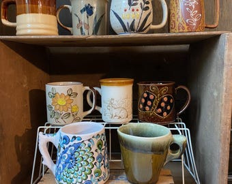 May include: A collection of eight ceramic mugs with various designs and colors. The mugs are displayed on a wooden shelf and a wire rack. The mugs feature floral, abstract, and geometric patterns, with earth-tone and vibrant color schemes.