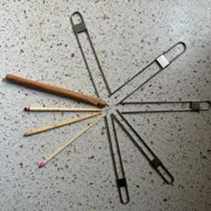 May include: Five silver-toned metal stitch holders arranged in a radial pattern, accompanied by one wooden pencil and four thin wooden sticks of varying lengths, all positioned to point toward a central convergence point on a speckled surface.