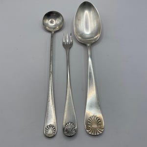 Puede incluir: Tres cubiertos de plata: un cucharón pequeño, un tenedor y una cuchara grande. Cada pieza tiene un diseño de concha decorativa en la base del mango. Los cubiertos están dispuestos verticalmente sobre un fondo gris.