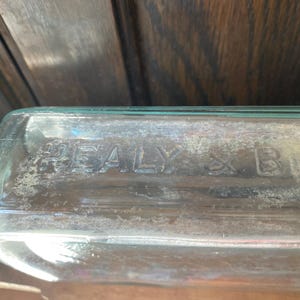 Puede incluir: Primer plano de una botella de vidrio transparente con el texto "HEALY & B" en relieve en el lateral.