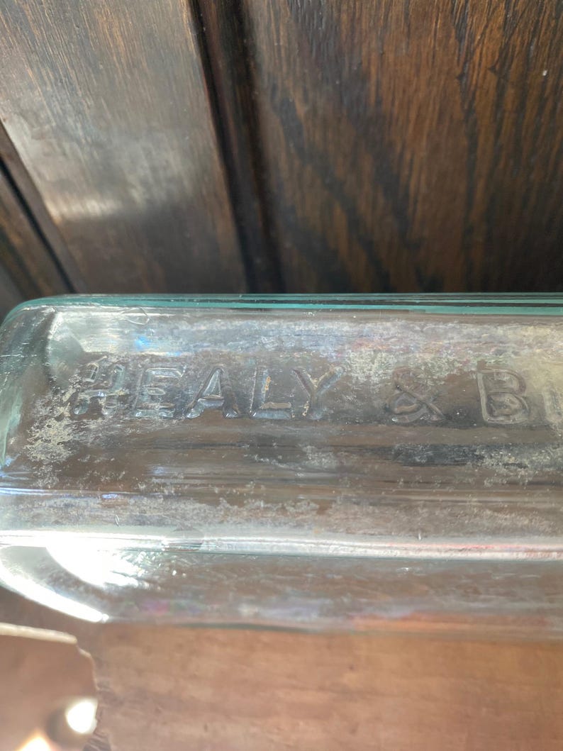 Puede incluir: Primer plano de una botella de vidrio transparente con el texto "HEALY & B" en relieve en el lateral.