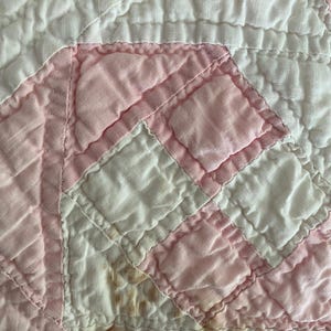 May include: A quilted textile featuring a geometric pattern of alternating white and light pink fabric squares. The fabric has a visible stitched grid texture throughout, with some small brown discoloration spots on the lower left side.