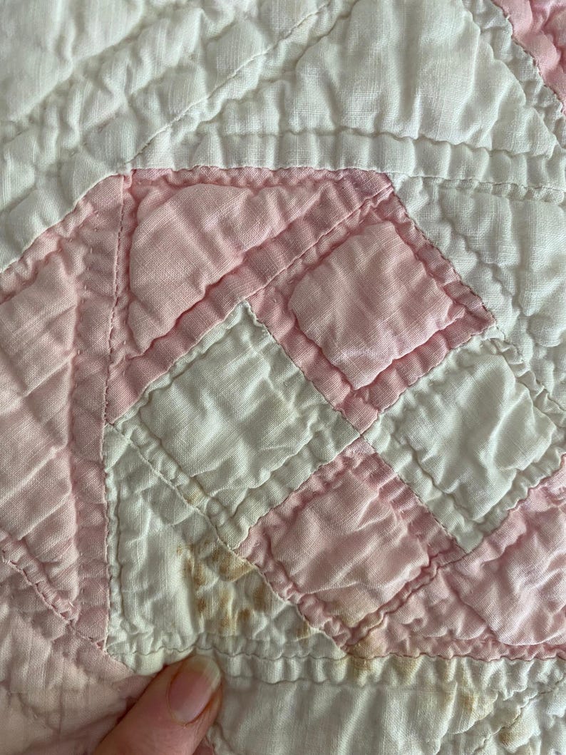 May include: A quilted textile featuring a geometric pattern of alternating white and light pink fabric squares. The fabric has a visible stitched grid texture throughout, with some small brown discoloration spots on the lower left side.