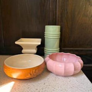 May include: A selection of ceramic bowls and vases in different shapes and colours. Features an orange bowl with a floral design, a pink scalloped bowl, a light green cylindrical vase, and a cream-coloured square vase. The items are displayed on a speckled countertop.