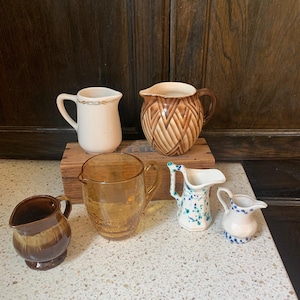 May include: A collection of six small jugs in various styles and colours. The jugs include a cream-coloured jug with a gold design, a brown and tan woven-patterned jug, and a clear amber glass jug.