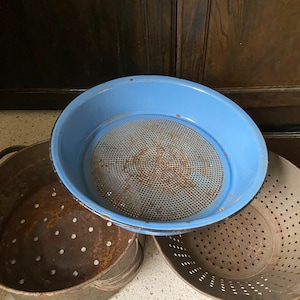 Puede incluir: Una colección de coladores y escurridores de cocina antiguos. El colador principal es de color azul claro con una malla metálica. Otros coladores son de metal con varios patrones de agujeros, mostrando signos de edad y uso. Los artículos están dispuestos sobre una superficie clara.