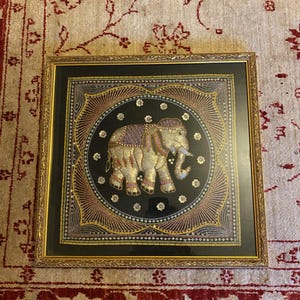 Op de afbeelding: Ingelijst kunstwerk met een sierlijk olifantenontwerp. De olifant is versierd met gouden en zilveren pailletten, tegen een zwarte achtergrond met een decoratieve rand. De lijst is goudkleurig.