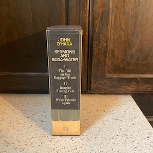 May include: A vintage paperback book titled "Sermons and Soda-Water" by John O'Hara. The book cover is black with yellow text and features three sections: "The Girl on the Baggage Truck", "Imagine Kissing Pete", and "We're Friends Again".