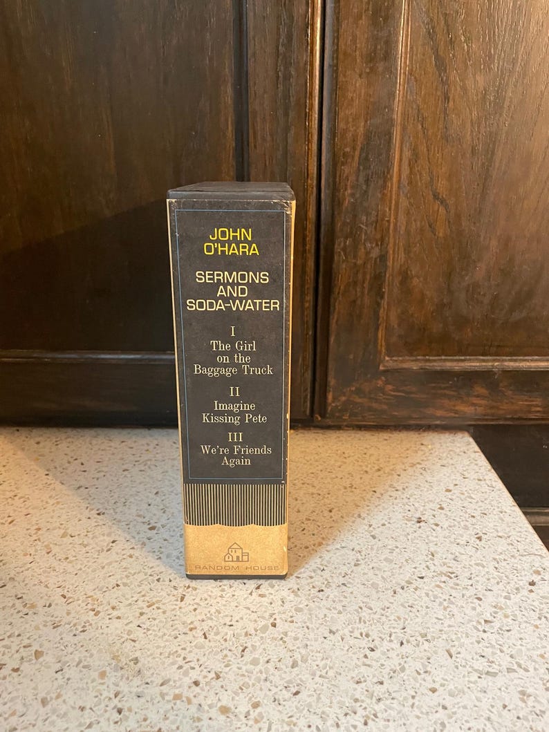 May include: A vintage paperback book titled "Sermons and Soda-Water" by John O'Hara. The book cover is black with yellow text and features three sections: "The Girl on the Baggage Truck", "Imagine Kissing Pete", and "We're Friends Again".