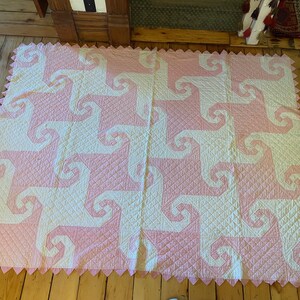 May include: A rectangular quilt featuring a repeating pattern of interlocking white and light pink spiral shapes. The fabric has a quilted diamond texture throughout, and the edges are finished with a decorative pink sawtooth border.